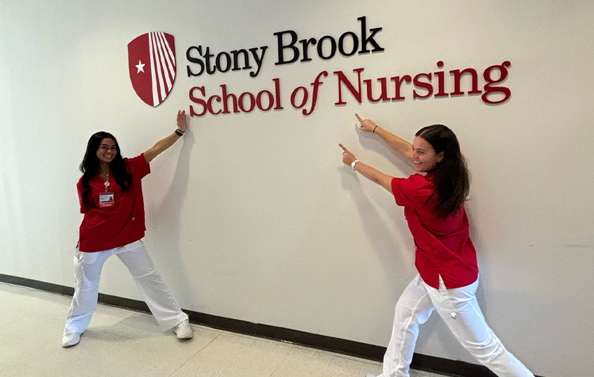 Two nursing students pointing at a Stony Brook School of Nursing sign. 