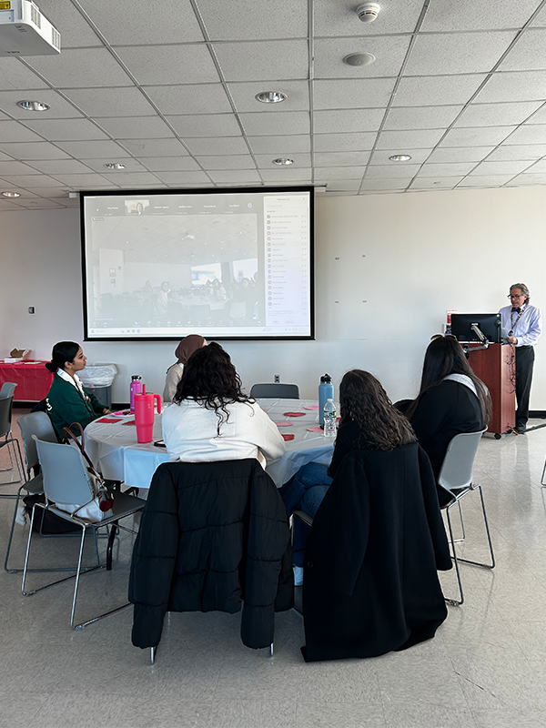 A group of people sitting at a table, a projector screen displaying a Zoom meeting room, and someone speaking at a podium.
